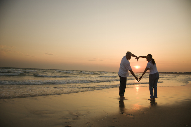 Mid-adult couple making heart shape with arms on beach at sunset.